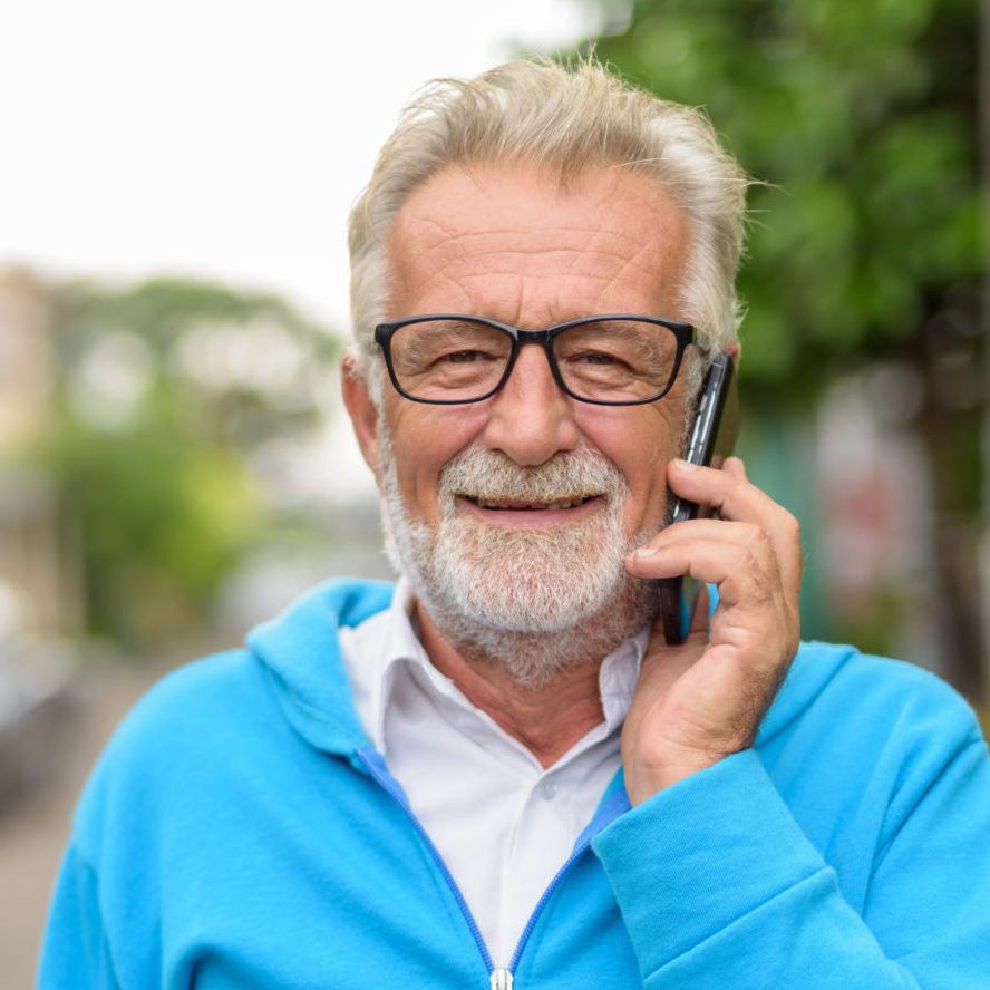 Happy handsome senior bearded man smiling while talking on mobile phone and wearing eyeglasses outdoors